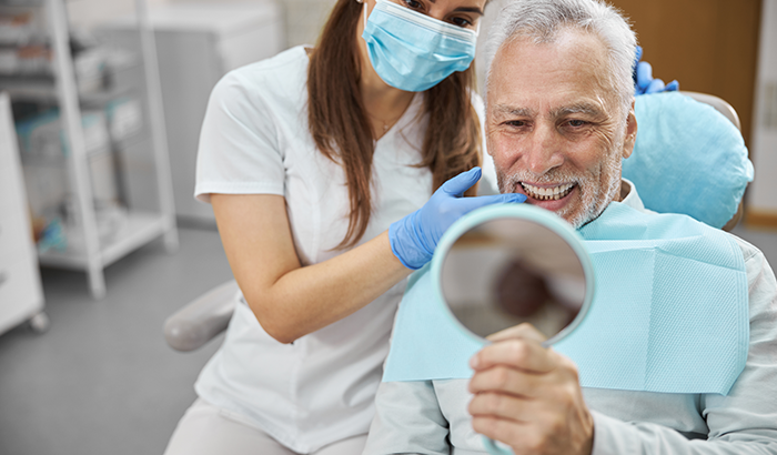 Older man in a dentist chair holding up a hand mirror and looking at his new dental implants at Peak Endodontics in Covington, WA